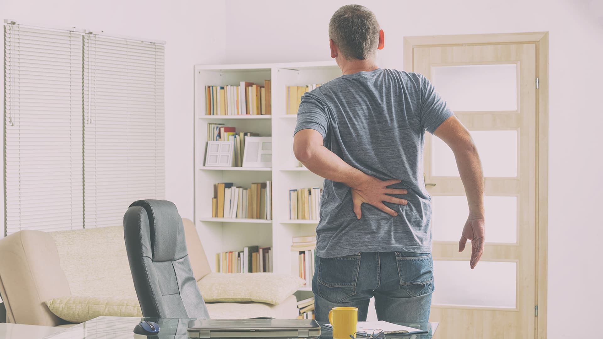 A man standing next to a desk with his hand on his lower back indicating pain