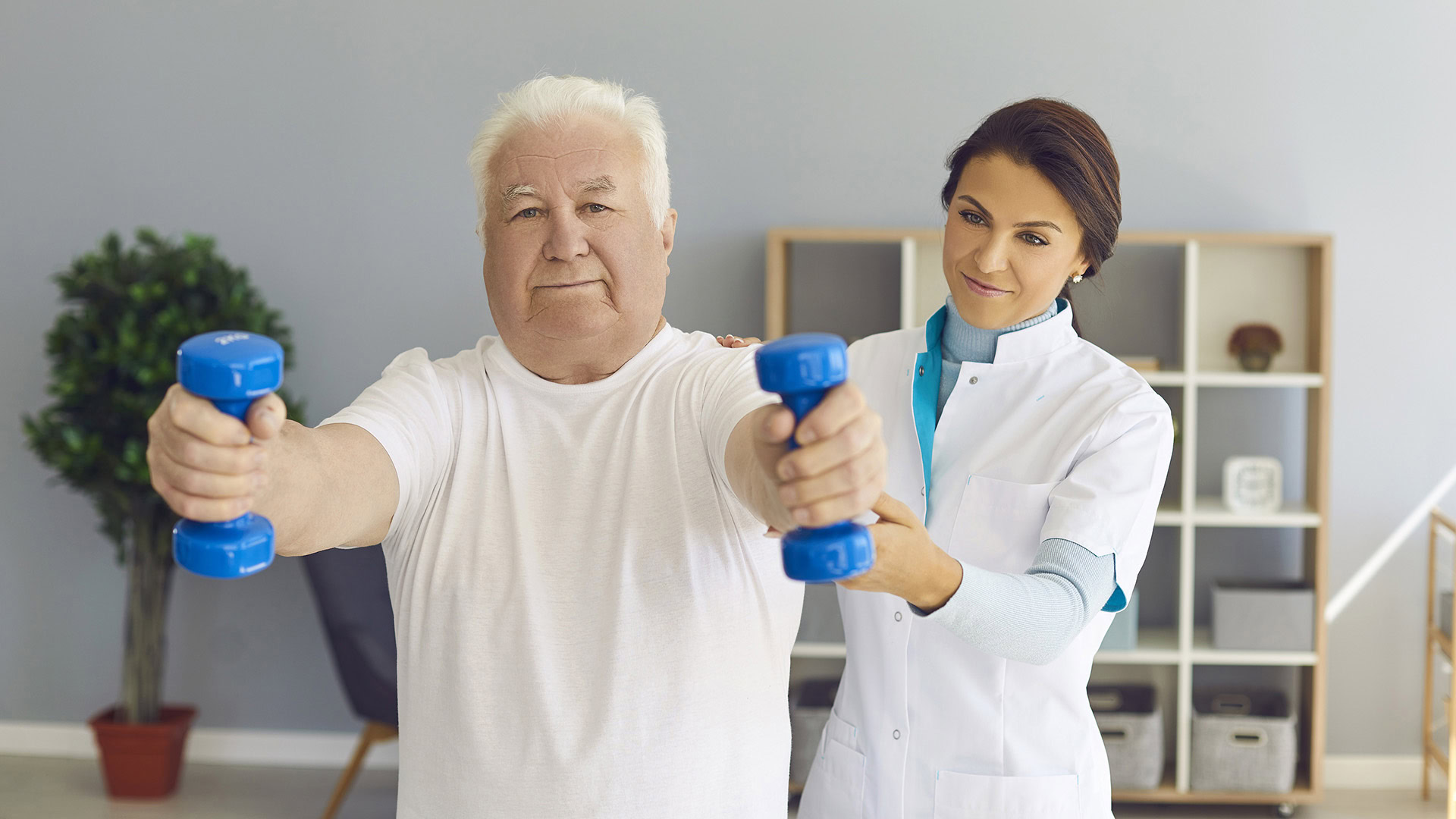 Doctor supports an old man's arms as he lifts dumbbells in medical office.